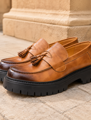 Mocassini uomo cognac con nappine e suola carrarmato nera su pavimentazione in pietra
