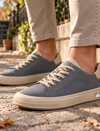 Sneakers uomo grigie con lacci chiari e suola a contrasto, indossate su pavimentazione in pietra